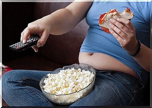 Girl Sitting On The Couch Watching TV With A Tub Of Popcorn On Her Lap And A Bun In Her Hand As An Example Of Bad Weight Loss Habits